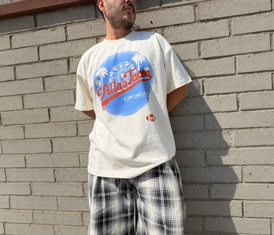 Cream Coachella souvenir T-shirt worn with plaid trousers against a brick wall