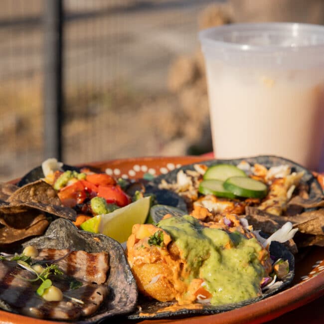 Blue-corn tacos with cheese skirts, guacamole, diced onion and cilantro, beside a drink